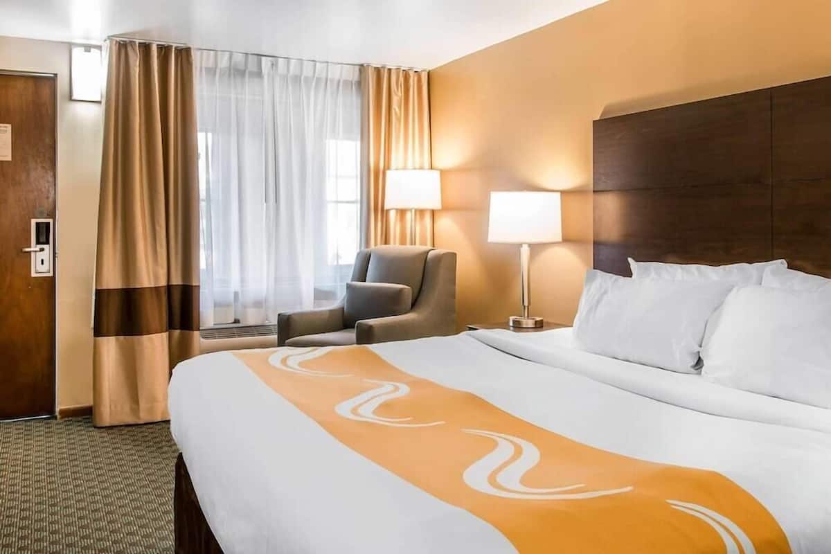 A hotel room with a large bed, a chair, a desk with a lamp, and a window with curtains.