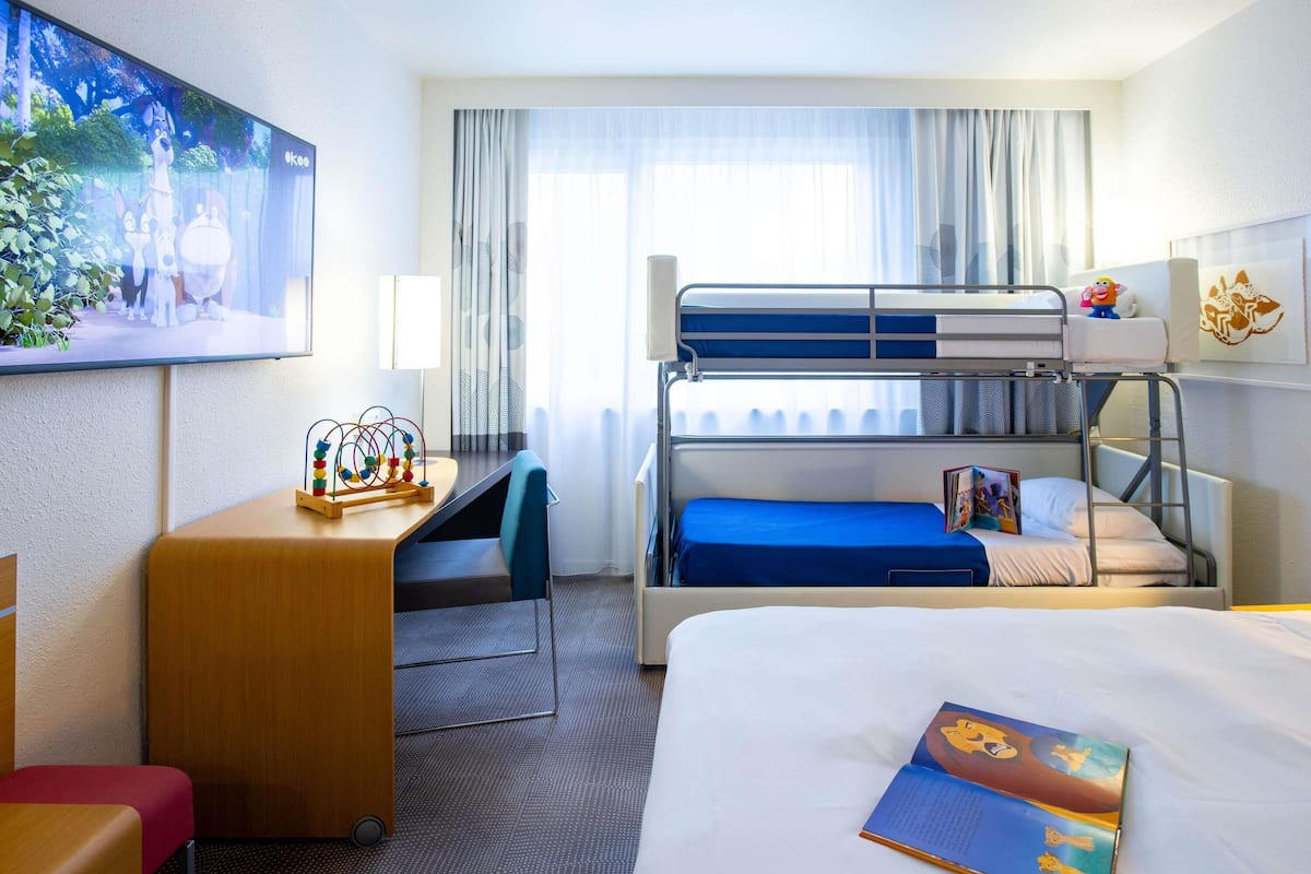 Une chambre d’hôtel avec des lits superposés, un bureau avec une lampe, une chaise, un lit avec un livre et un tableau au mur.