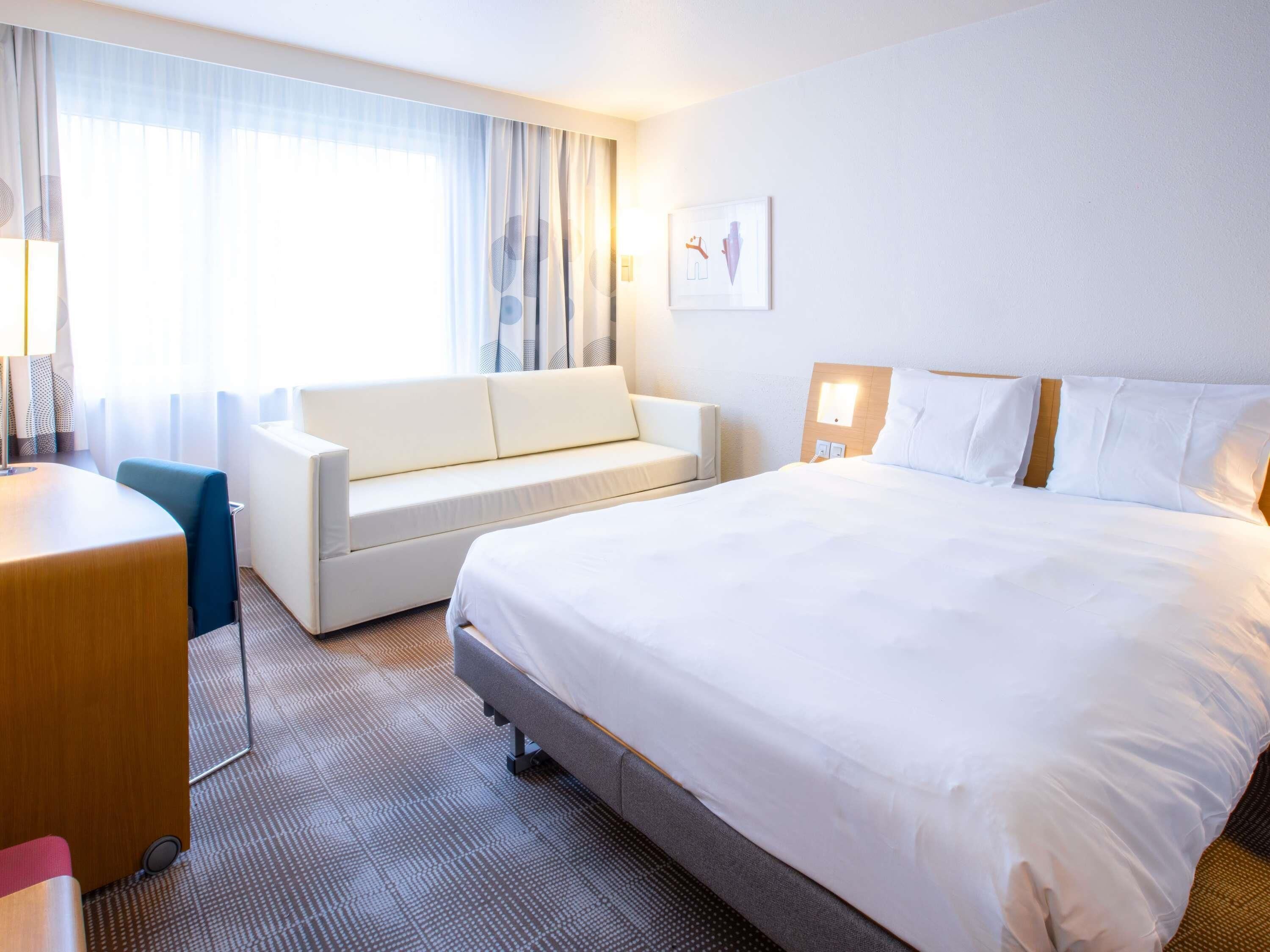 A hotel room with a bed, a sofa, a desk, and a window with curtains.