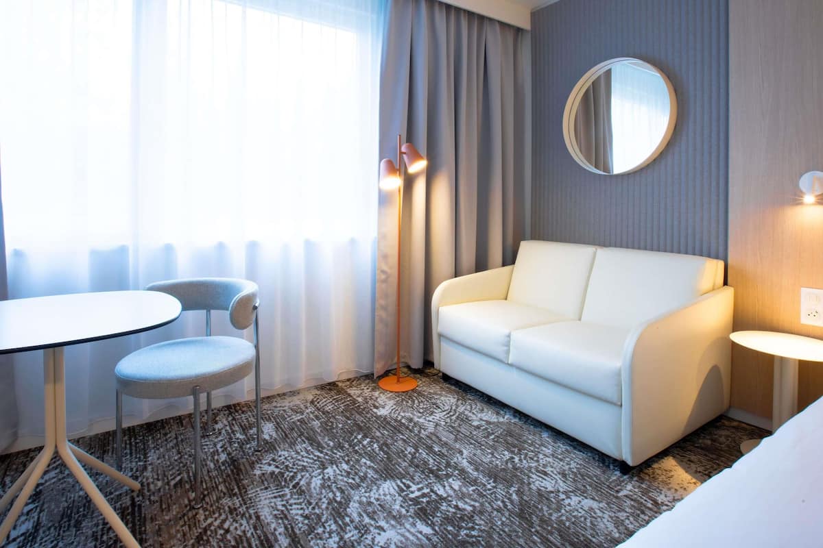 Une chambre d’hôtel avec un canapé blanc, un miroir rond, une petite table et une chaise.