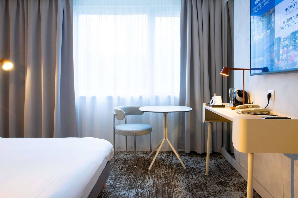 A hotel room with a bed, a desk with a lamp, a chair, and a large window with curtains.