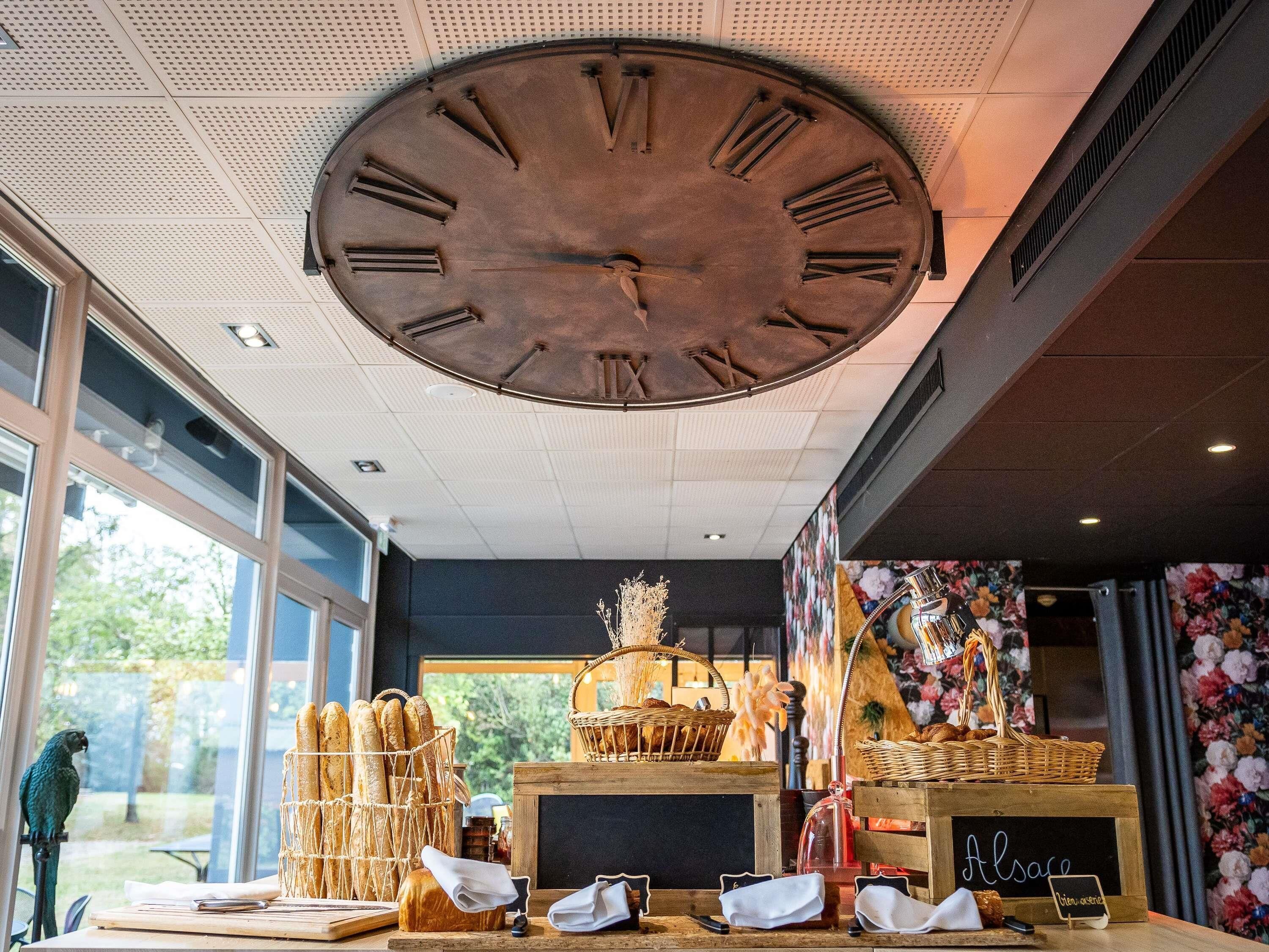 Un reloj de pared grande, un mostrador de madera con cestas de pan y una pizarra con la palabra 'Alsace'.