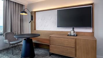 Une chambre d’hôtel dotée d’un grand téléviseur à écran plat fixé au mur, d’une table ronde avec une chaise, d’une commode en bois avec des tiroirs et d’une fenêtre avec des rideaux.
