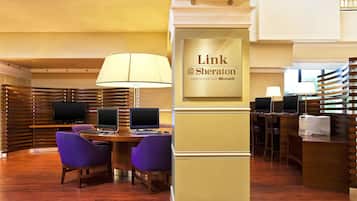 A hotel reception area with a reception desk, two computer stations, purple chairs, a large lamp, and a sign reading "Link @Sheraton experienced with Microsoft".