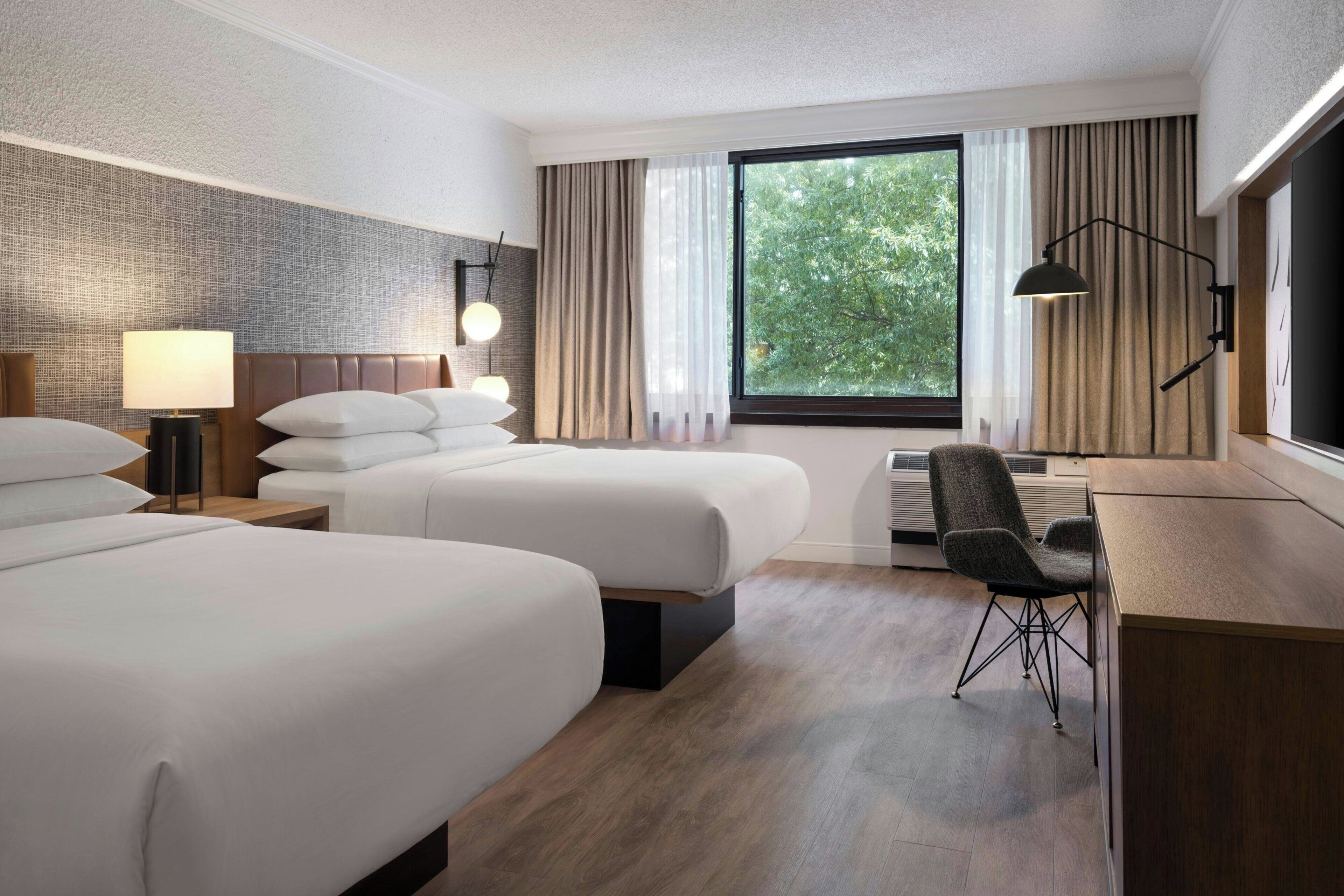 Habitación de hotel con dos camas, un escritorio, una silla, una lámpara y vistas al verde exterior.