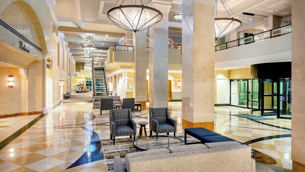 A spacious lobby with high ceilings, large columns, and a checkered floor pattern. There are seating areas with armchairs and a sofa, and a staircase in the background. - Sheraton Pentagon City Hotel (Arlington)