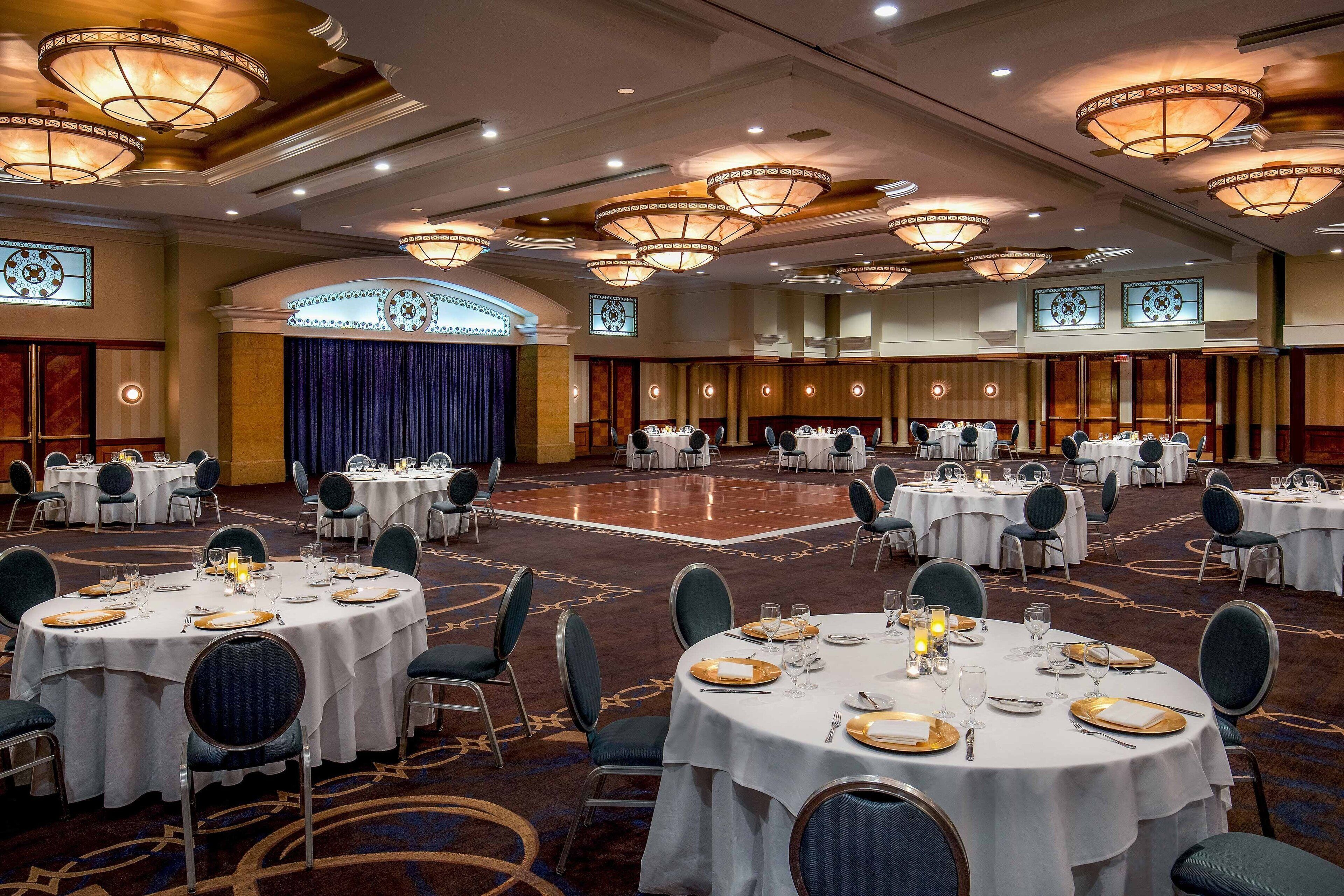 A banquet hall with round tables set for dining, featuring chandeliers and a carpeted floor.