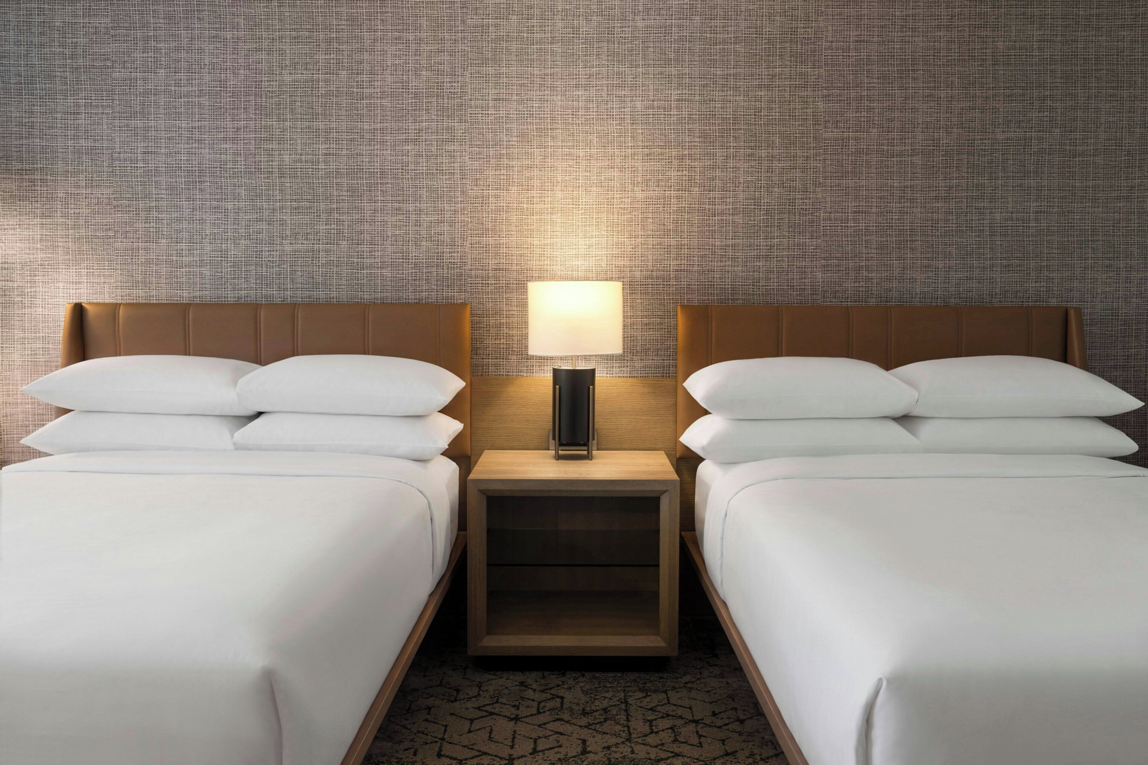 Habitación de hotel con dos camas, una mesita de noche con lámpara y una pared con textura.