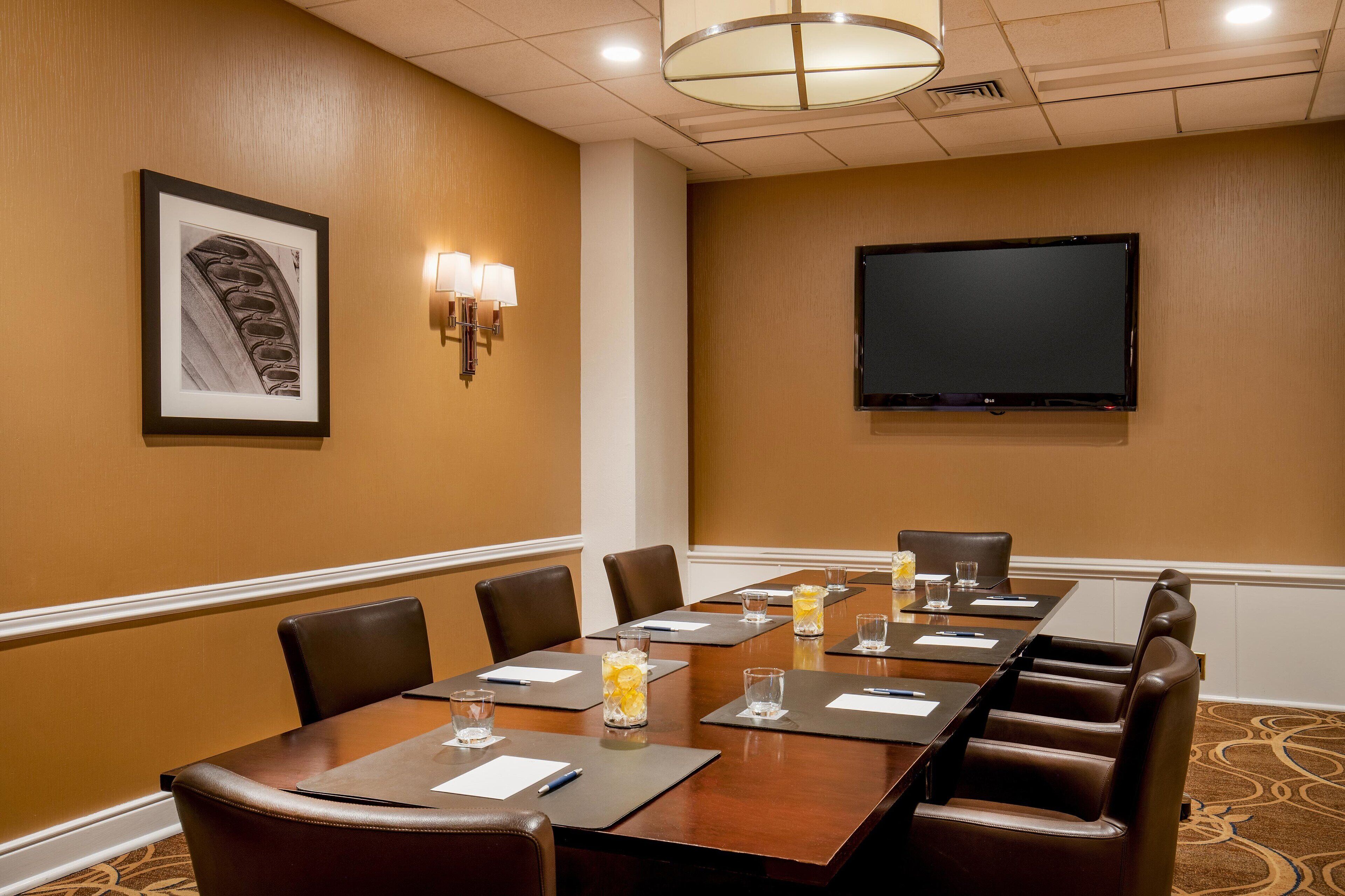 A conference room with a long table, chairs, a flat-screen TV, framed artwork, and wall sconces.