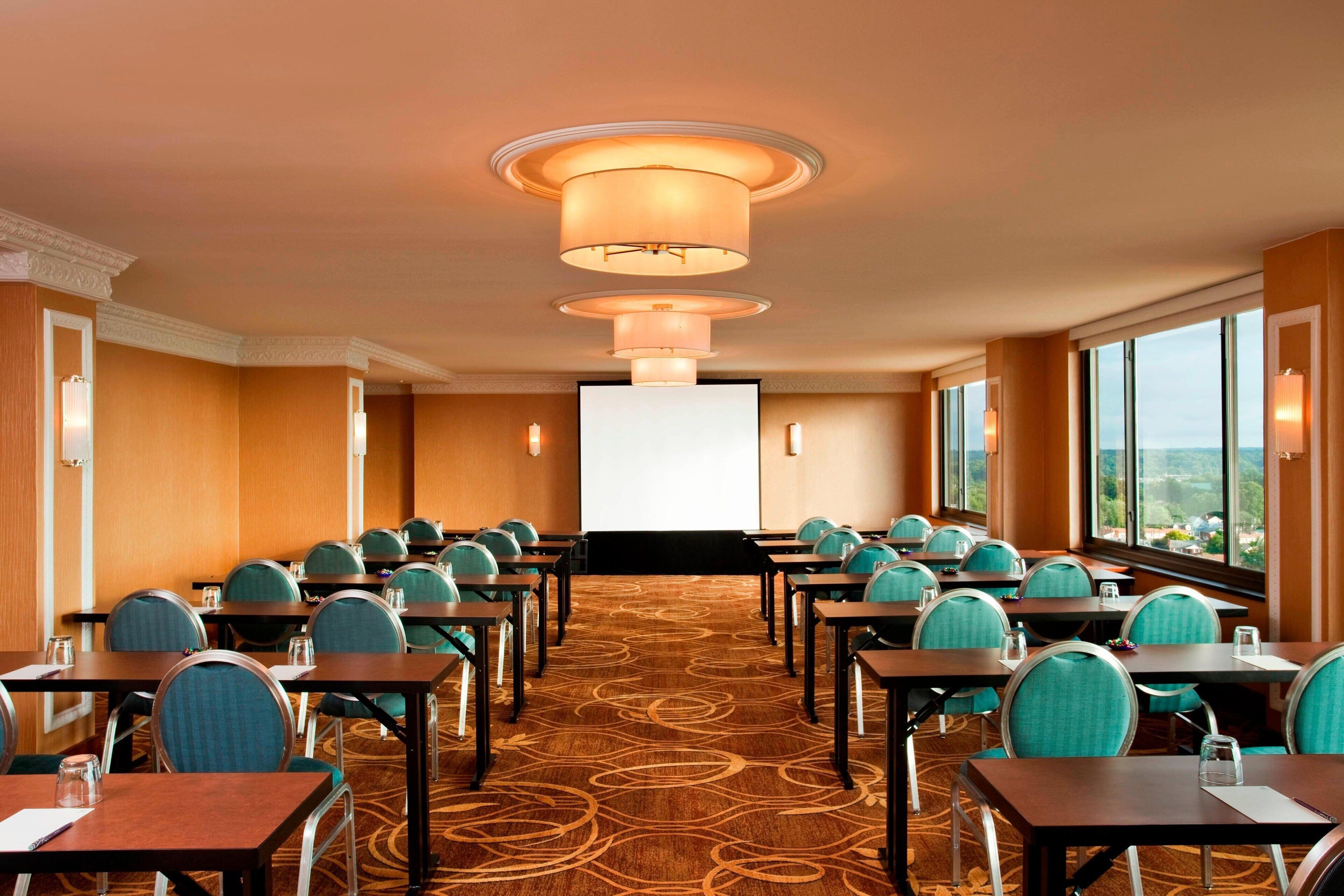 Une salle de conférence avec des rangées de tables et de chaises, un grand écran à l’avant et de grandes fenêtres offrant une vue dégagée.