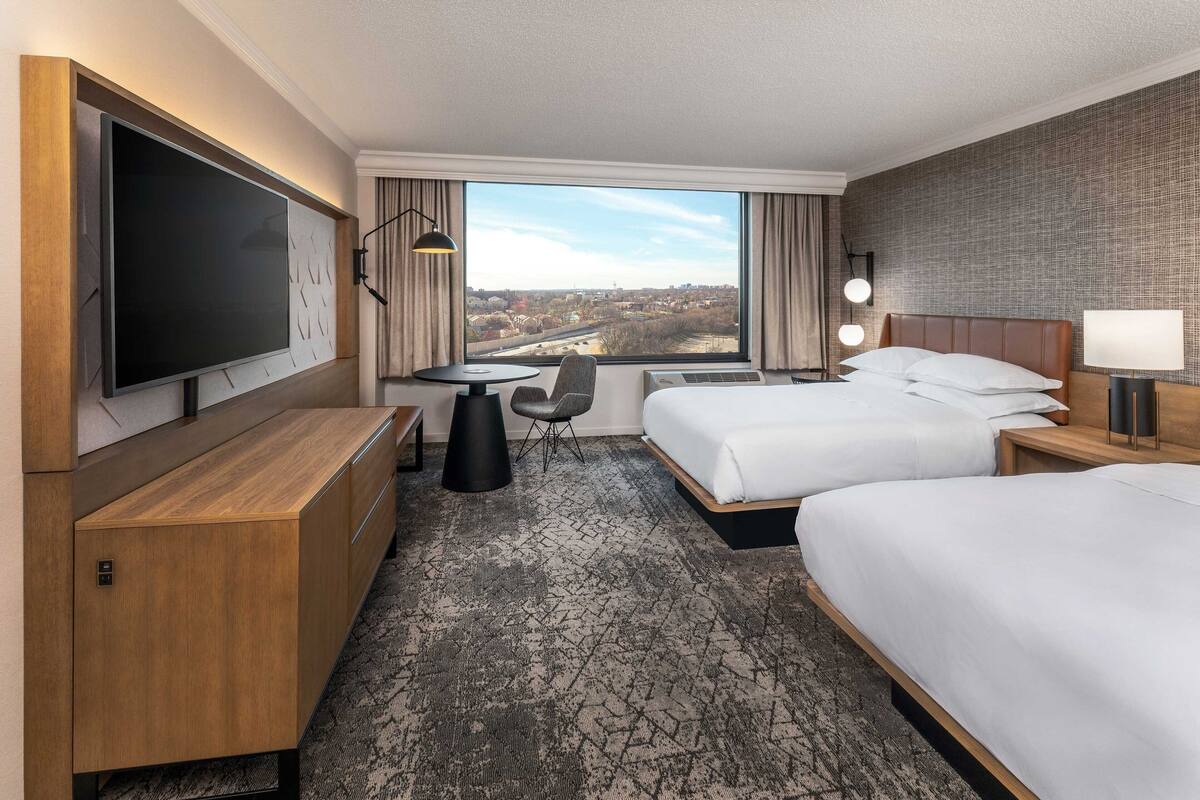 Hotellihuone, jossa on kaksi sänkyä, televisio, työpöytä ja suuri ikkuna, josta avautuu näkymä kaupunkiin.