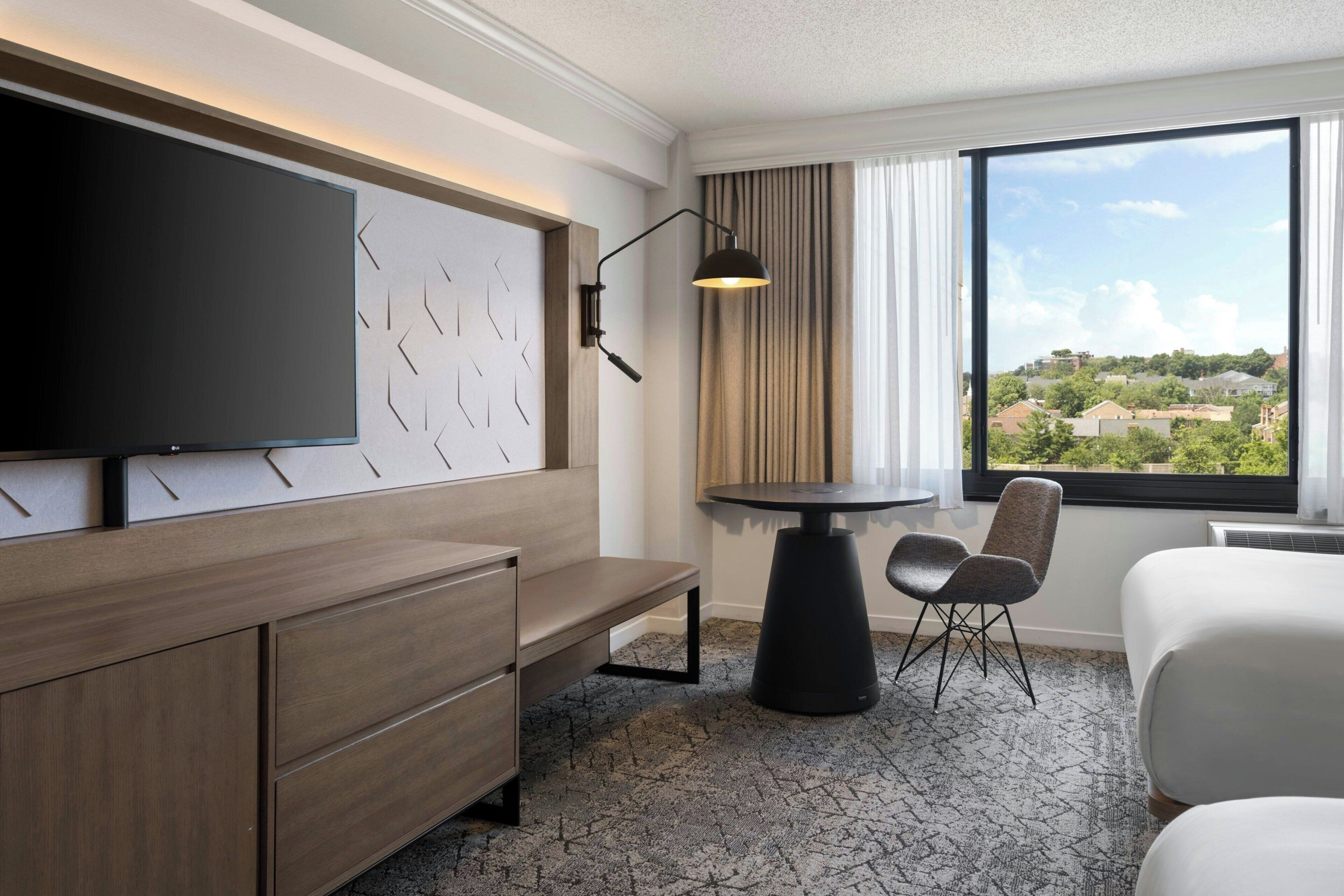 Et hotelværelse med et stort fladskærms-tv, et skrivebord med stol, et lille rundt bord og udsigt til et boligområde gennem et stort vindue.