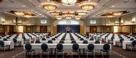 A conference room with rows of tables and chairs, a stage with a blue curtain, and ornate chandeliers.