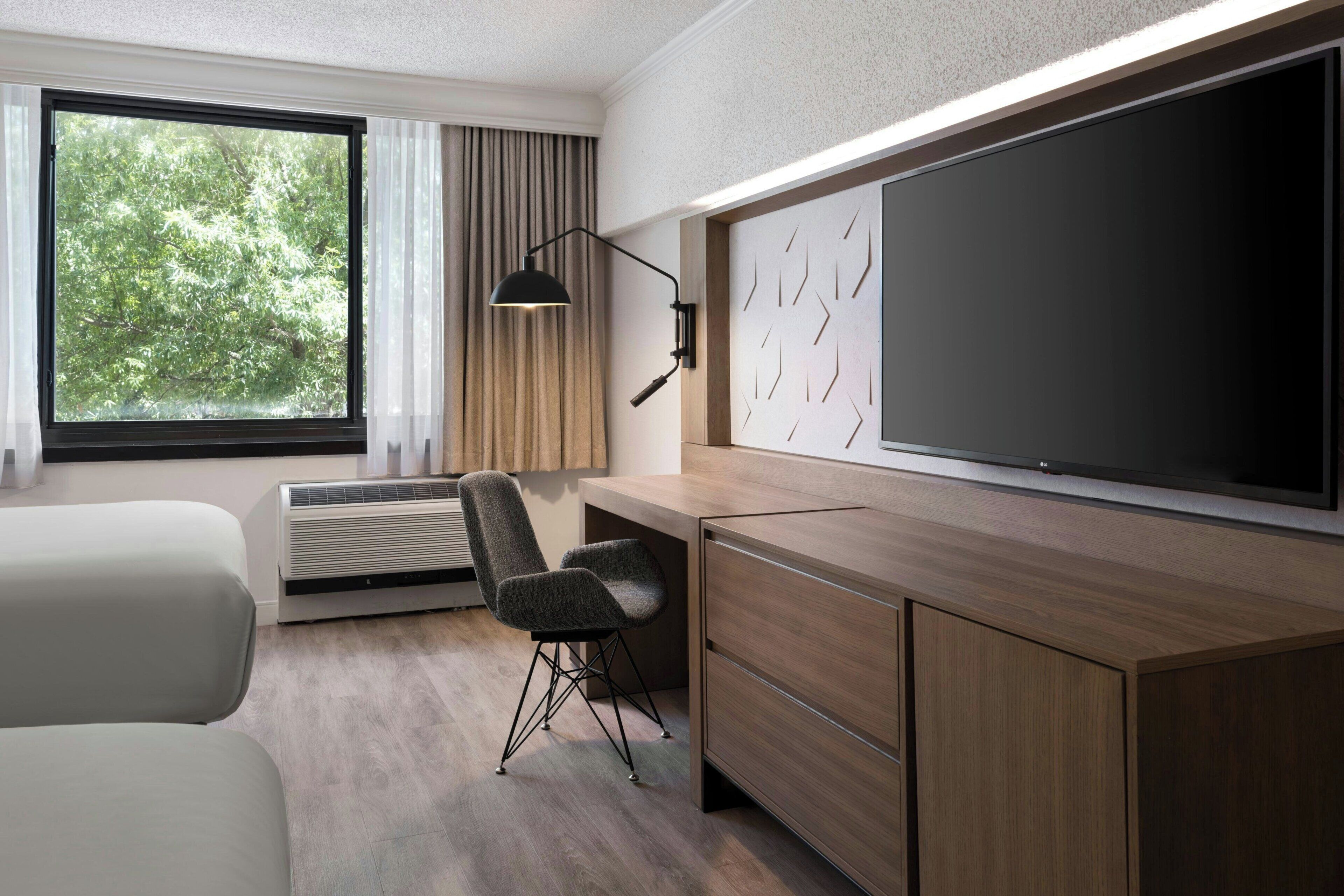 Una habitación de hotel moderna con un ventanal grande, un escritorio con silla, un televisor de pantalla plana y un aire acondicionado instalado en la pared.