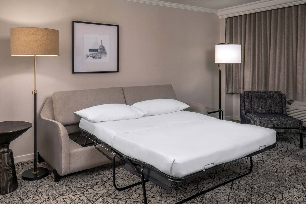 Hotellihuone, jossa on sänky, tuoli, lattiavalaisin, pöytä ja seinällä oleva kehystetty kuva.