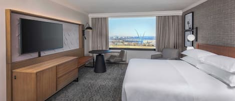 A hotel room with a large bed, a flat-screen TV, a wooden bench, a small table, and a view of the city.