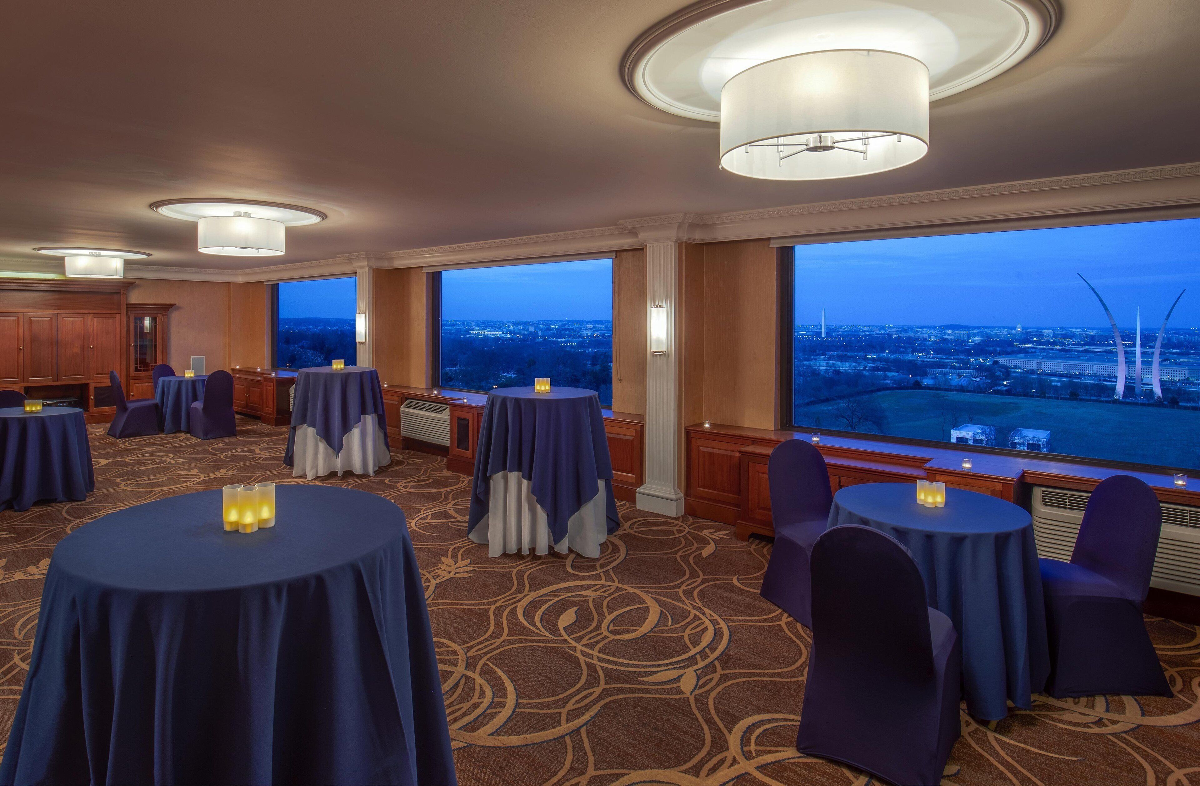 A hotel room with large windows, round tables, and chairs, and a view of a cityscape at dusk.