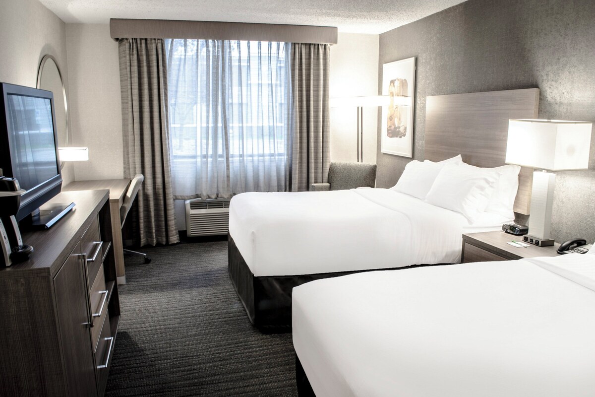 Ein Hotelzimmer mit zwei Betten, einem Schreibtisch mit Fernseher und einem großen Fenster mit Vorhängen.