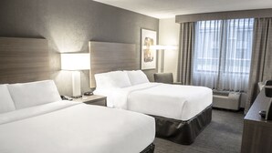 Quarto de hotel com duas camas, uma escrivaninha, uma cadeira, um abajur e uma janela grande com cortinas.