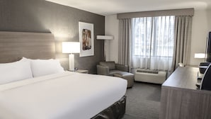 Quarto de hotel com uma cama grande, uma escrivaninha, uma cadeira, um sofá, um abajur e uma janela com cortinas.
