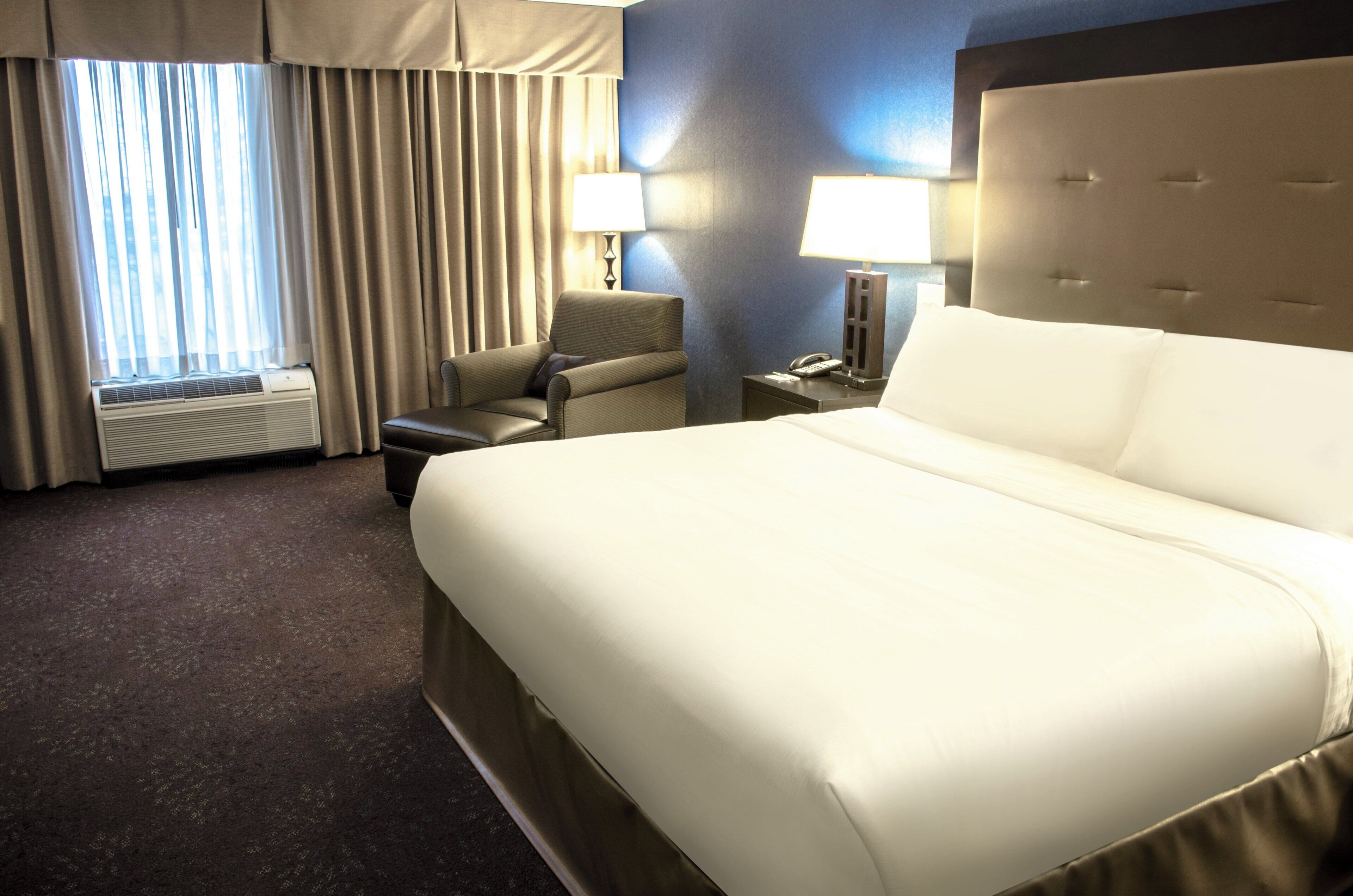 A hotel room with a large bed, a chair, a desk with a lamp, and a window with curtains.