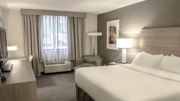 Quarto de hotel com uma cama grande, uma escrivaninha com computador, uma cadeira, um abajur e uma janela com cortinas.