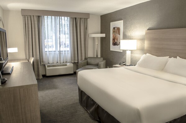 Ein Hotelzimmer mit einem großen Bett, einem Schreibtisch mit Computer, einem Sessel, einer Lampe und einem Fenster mit Vorhängen.
