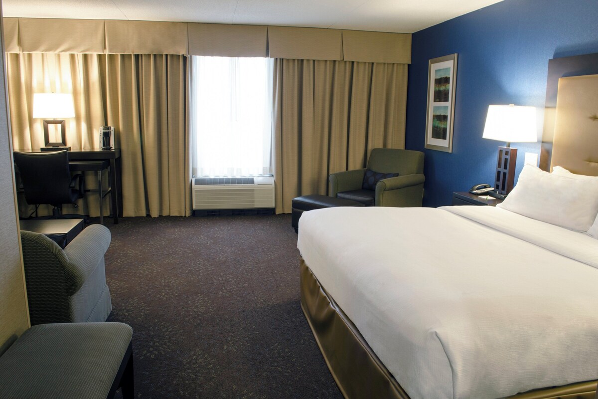 Ein Hotelzimmer mit Bett, Schreibtisch, Stuhl, Lampe und einem fenster mit Vorhängen.