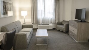 Quarto de hotel com sofá, mesa de centro, televisão, luminária e janela com cortinas.