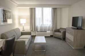 Ein Hotelzimmer mit einer Couch, einem Couchtisch, einem Fernseher, einer Lampe und einem Fenster mit Vorhängen.