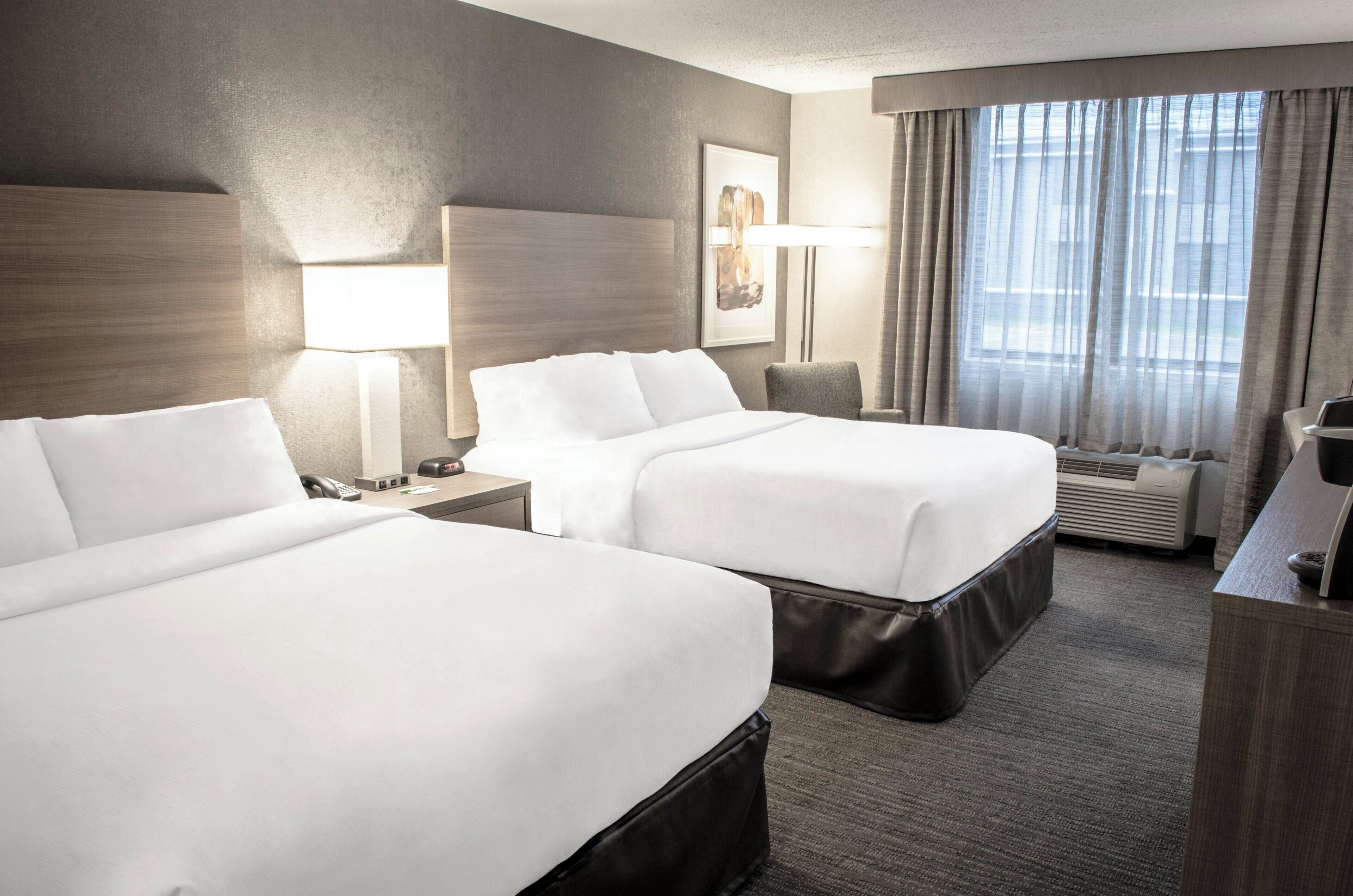 A hotel room with two beds, a desk, a chair, a lamp, and a large window with curtains.
