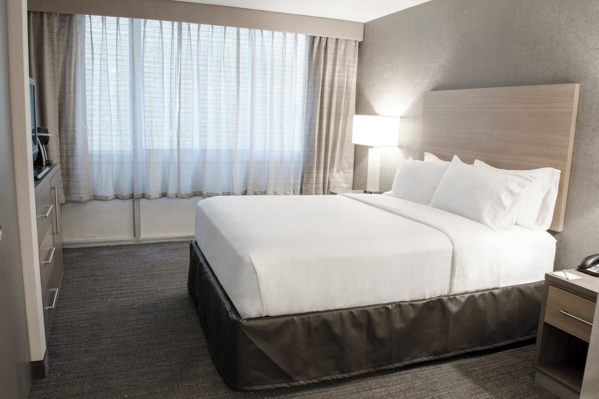 Ein Hotelzimmer mit einem großen Bett, einem Nachttisch mit Lampe, einem Schreibtisch mit Computer und einem Fenster mit Vorhängen.