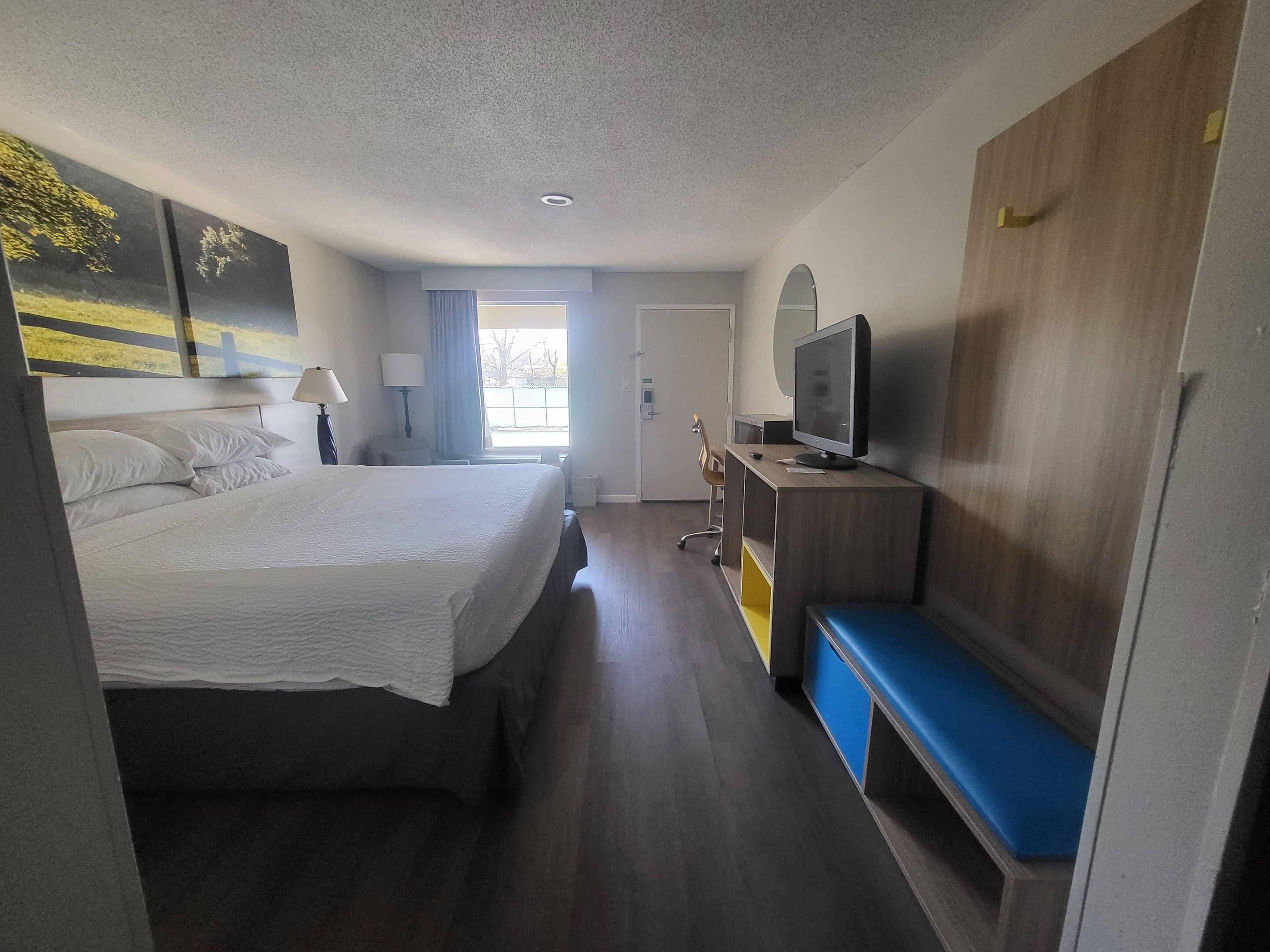 A hotel room with a bed, a desk with a computer and TV, a bench, and a large window.