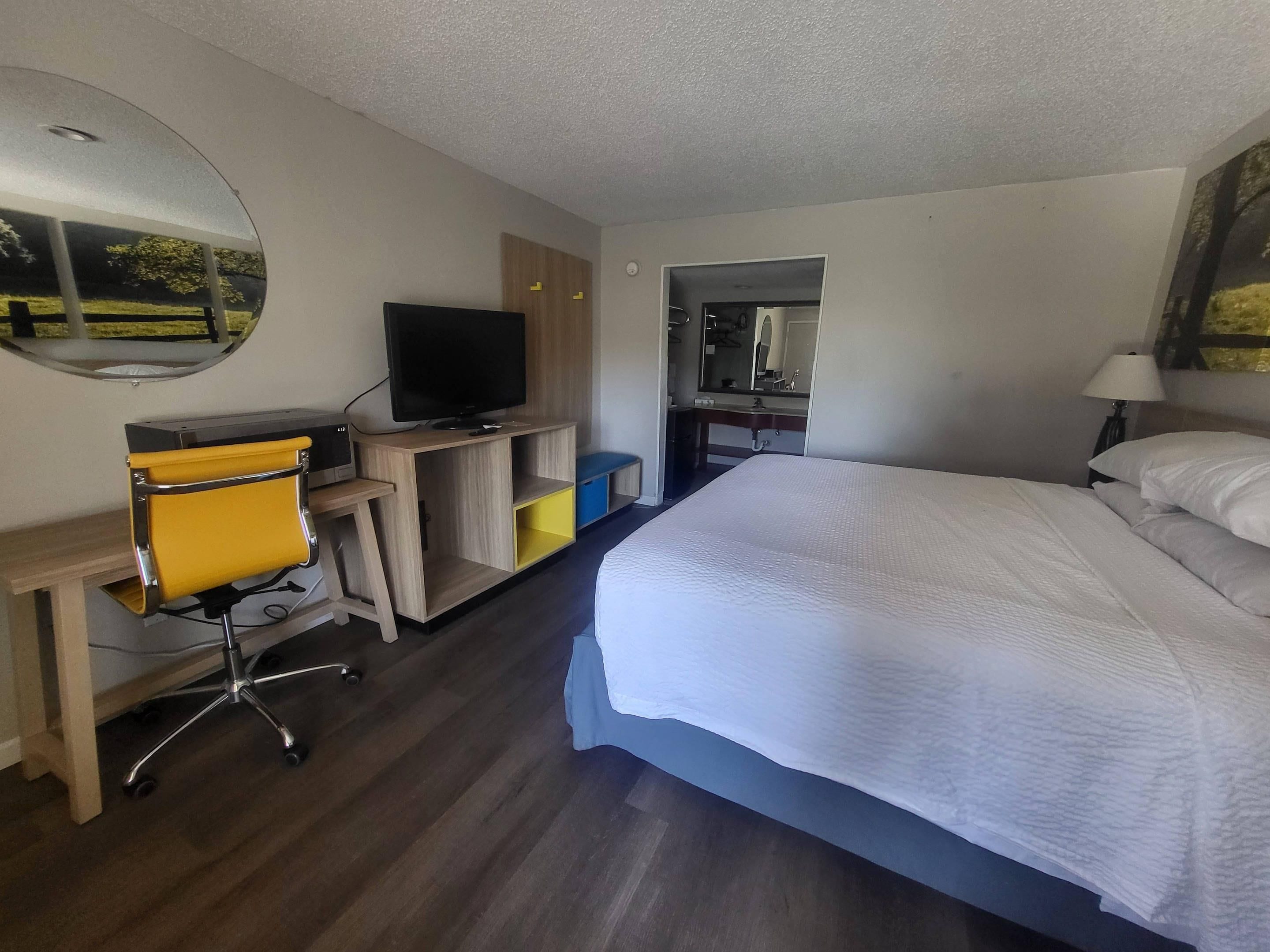 A hotel room with a bed, a desk with a chair, a television, a mirror, and a bathroom visible through an open door.