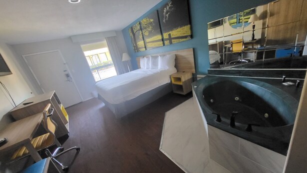 A modern hotel room with a bed, a small bedside table, a large bathtub, and a view of the outdoors. - Park Inn by Radisson, Attalla-Gadsden (Attalla)