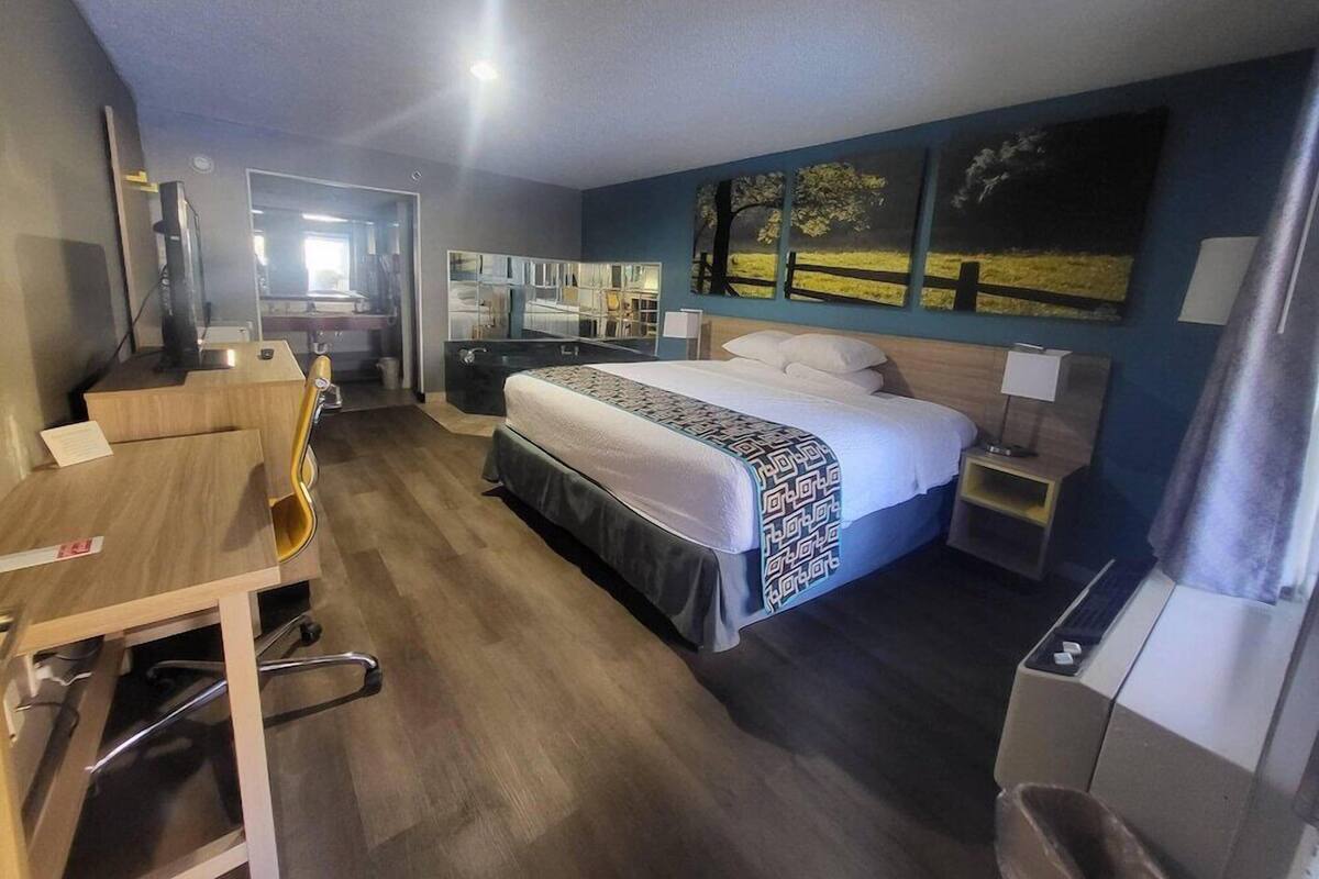 A hotel room with a bed, a desk, a chair, a mirror, and artwork on the wall.