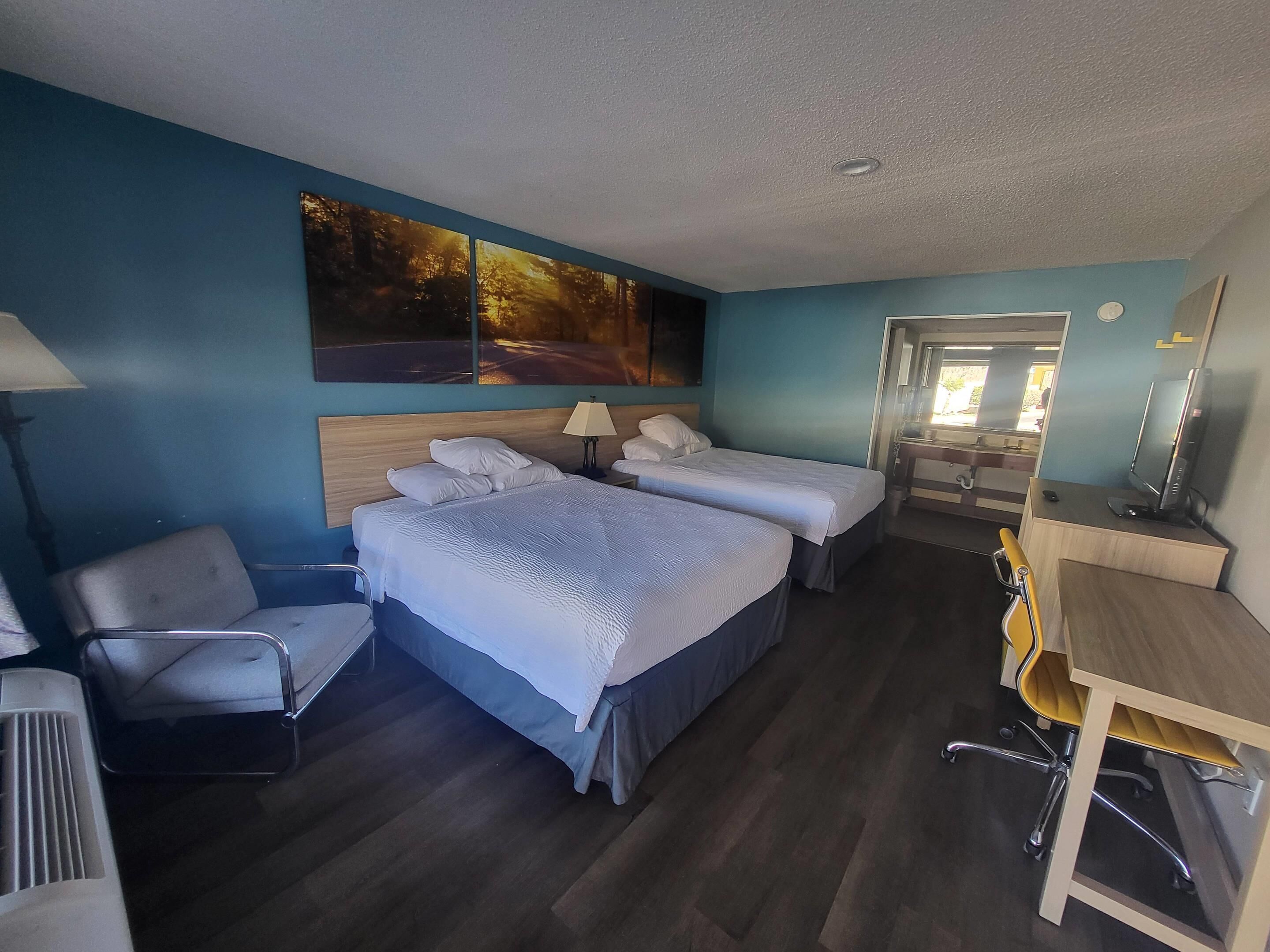 A hotel room with two beds, a desk, a chair, a lamp, and a painting on the wall.