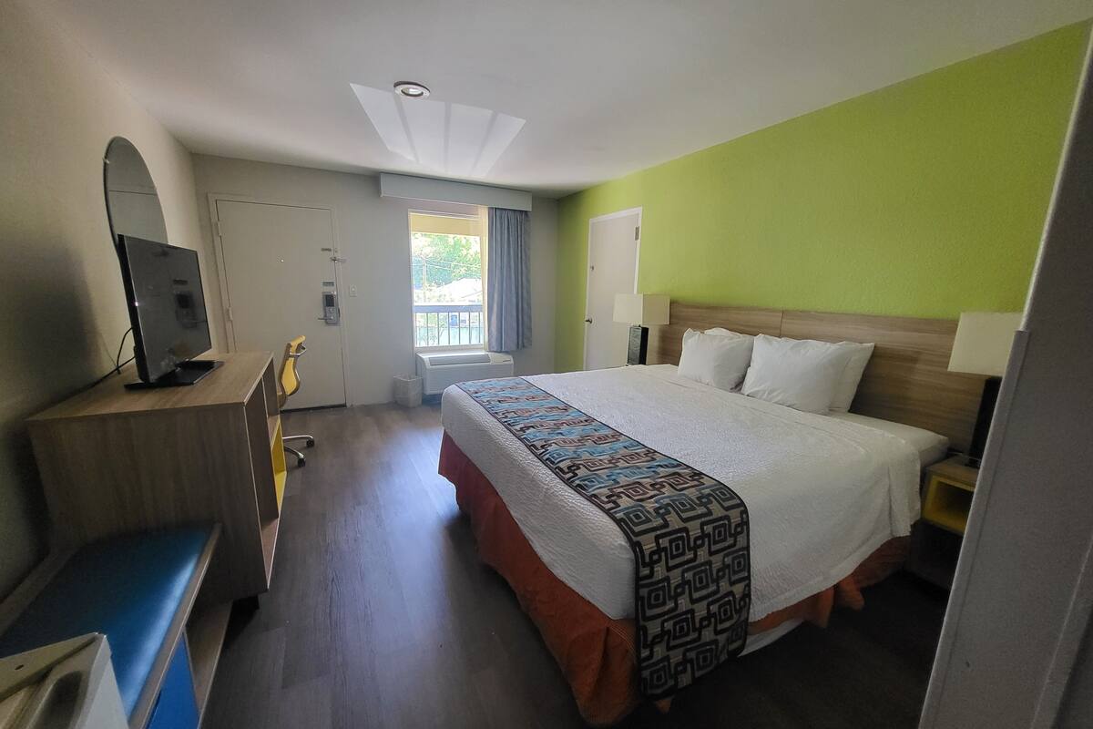 A hotel room with a bed, a desk with a computer, a television, a window with curtains, and a ceiling light.
