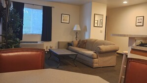 Habitación de hotel con kitchenette, sofá, mesa de comedor y un ventanal grande con persianas.