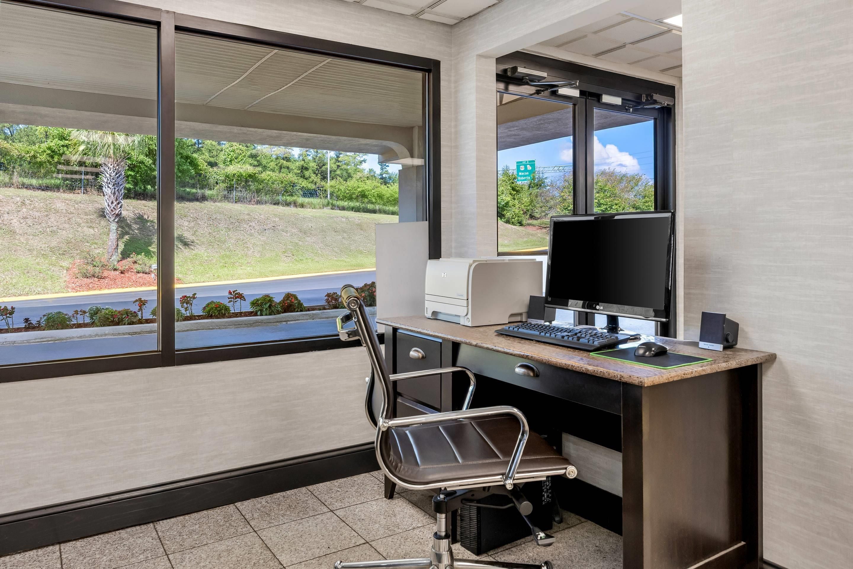 A modern office space with a desk, chair, computer, and large windows overlooking a landscaped area.