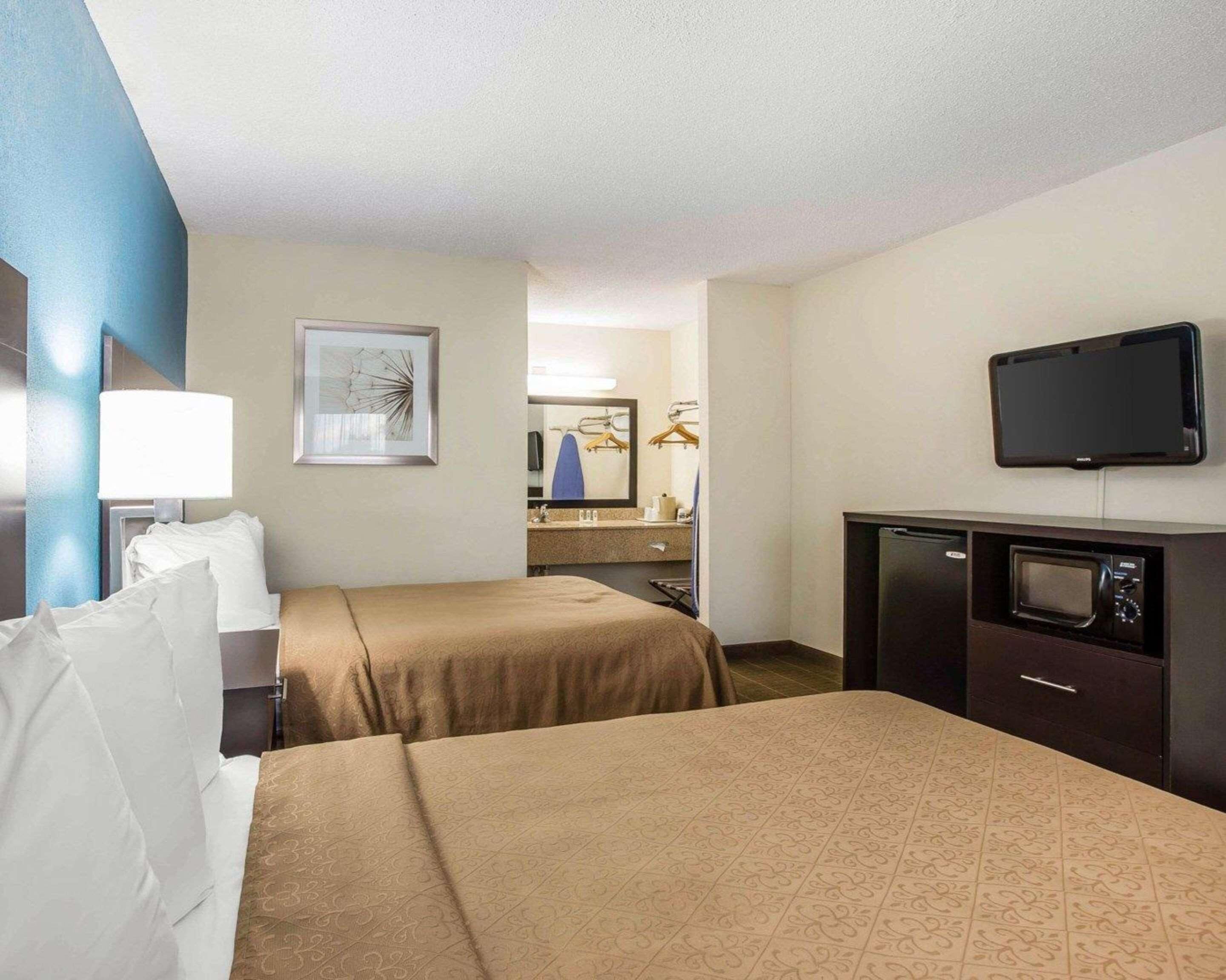 Habitación de hotel con dos camas, un escritorio con televisor, un espejo y un baño visible al fondo.