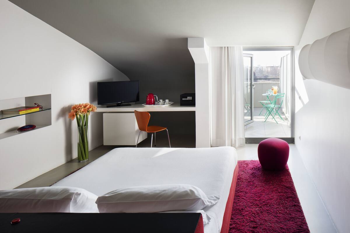 Una habitación de hotel moderna con cama, escritorio con televisor, silla, mesita con tetera roja y balcón con mesa y sillas.