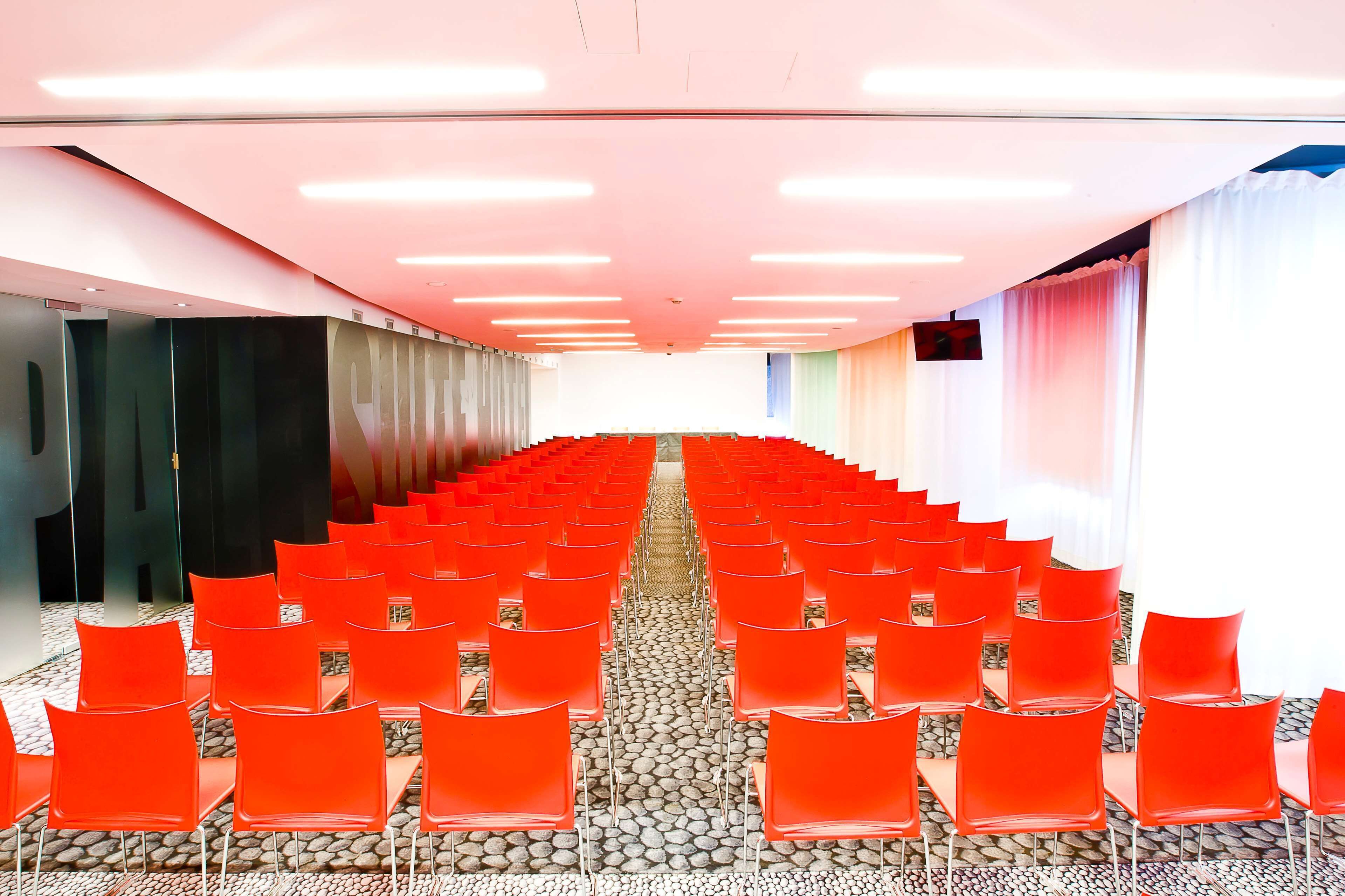 A conference room with rows of red chairs, a stone-patterned floor, and a bright ceiling.