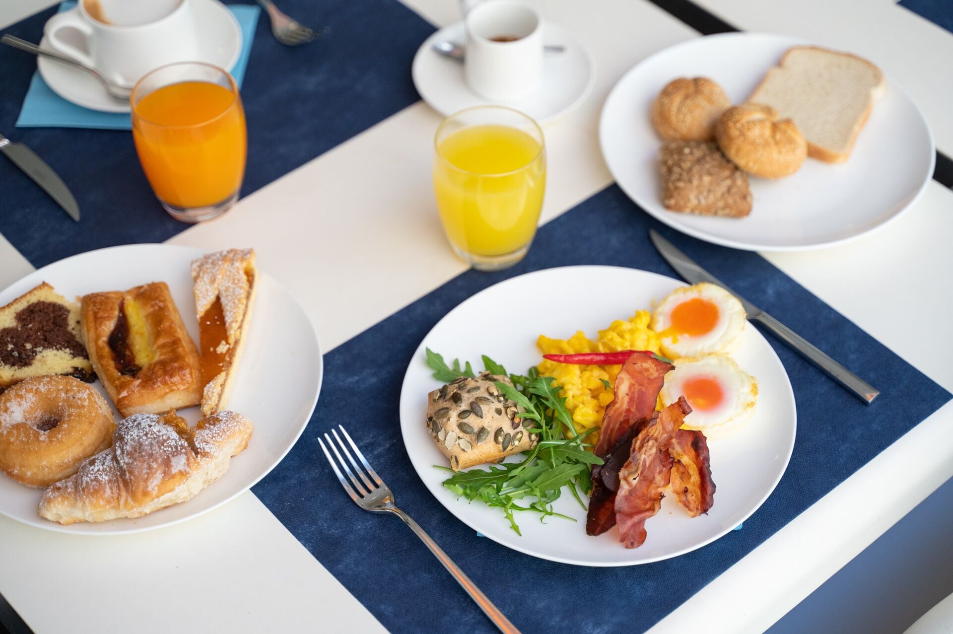 A breakfast table with plates of food, including pastries, eggs, bacon, and vegetables, along with cups of coffee and orange juice.