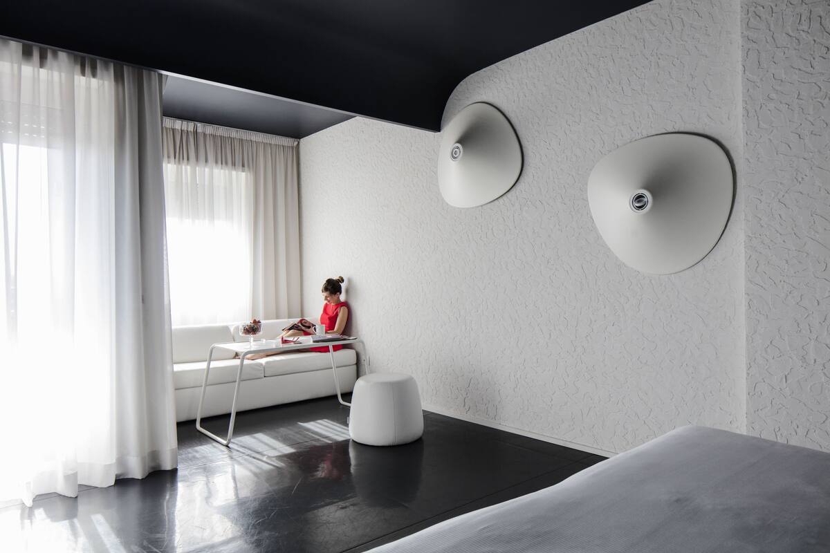 Una habitación de hotel moderna con un ventanal grande, un sofá blanco, una mesita pequeña y un reposapiés blanco.