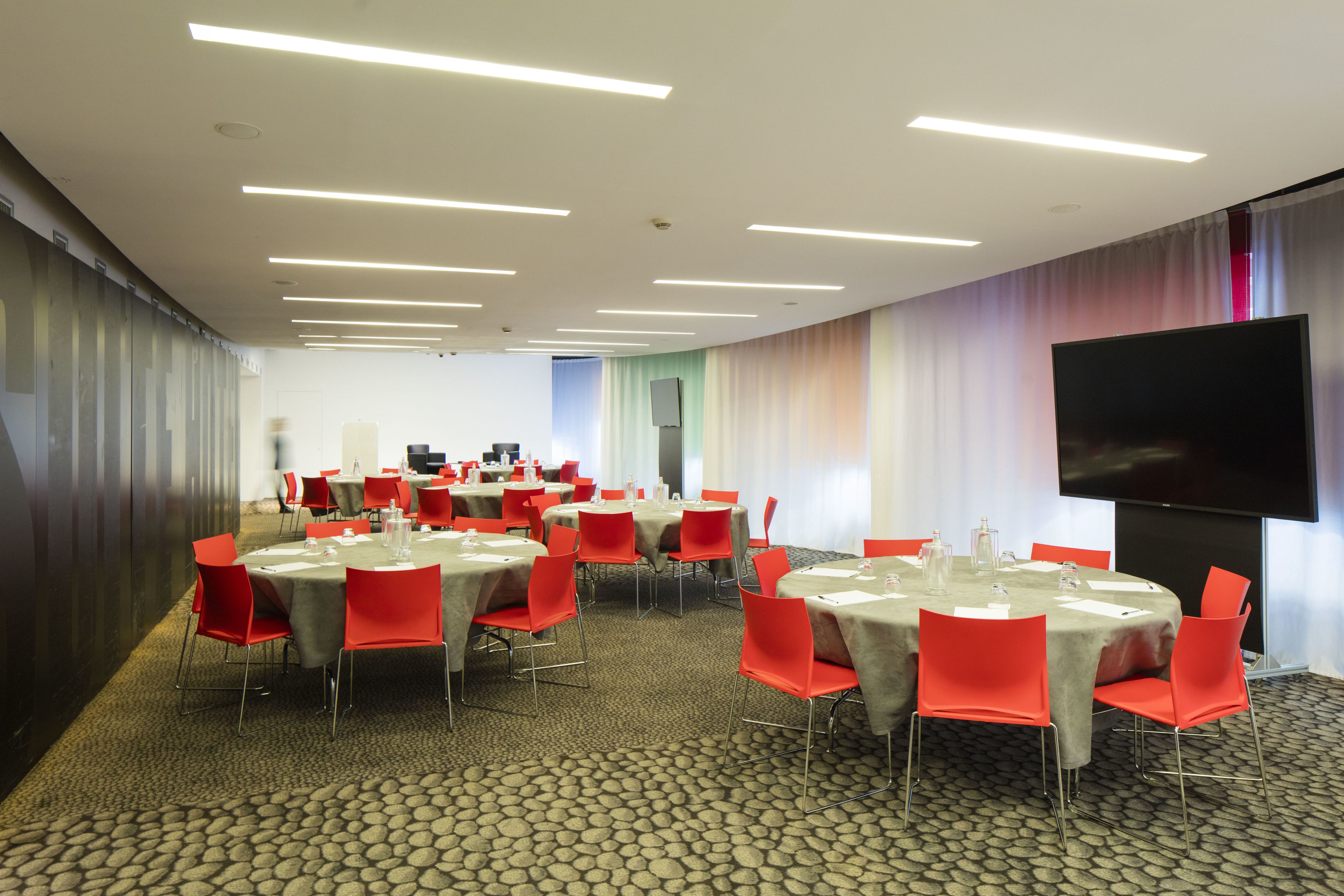 A conference room with multiple tables, red chairs, and a large flat-screen TV.