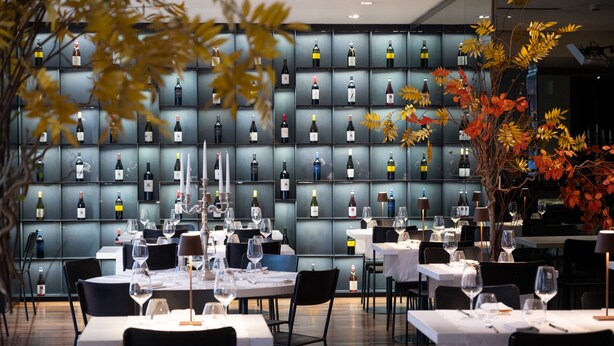 Un restaurante con mesas dispuestas para comer, una pared de botellas de vino y decoración con follaje otoñal.