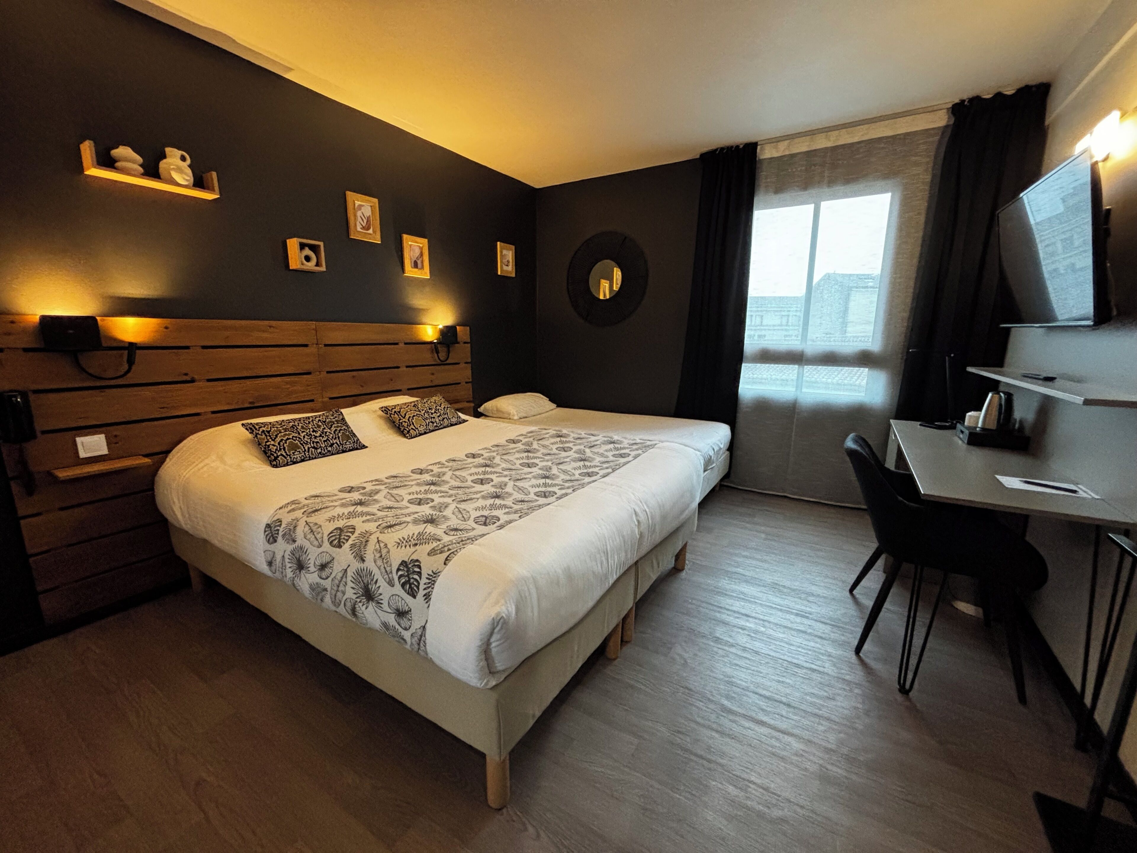 A modern hotel room with a wooden headboard, a large bed with patterned bedding, a desk with a chair, and a view of the city through the window.