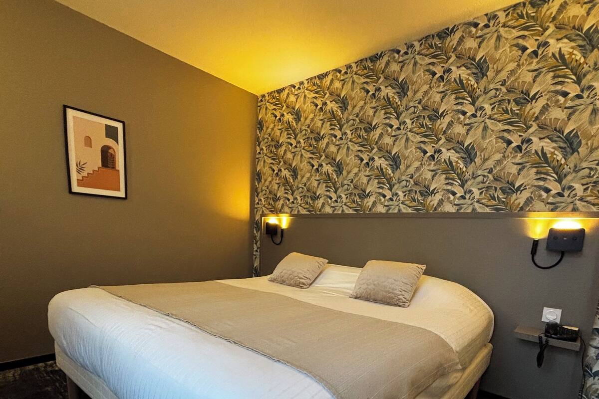 A hotel room with a large bed, two bedside lamps, a floral wallpaper headboard, a framed picture on the wall, and a dark wooden floor.