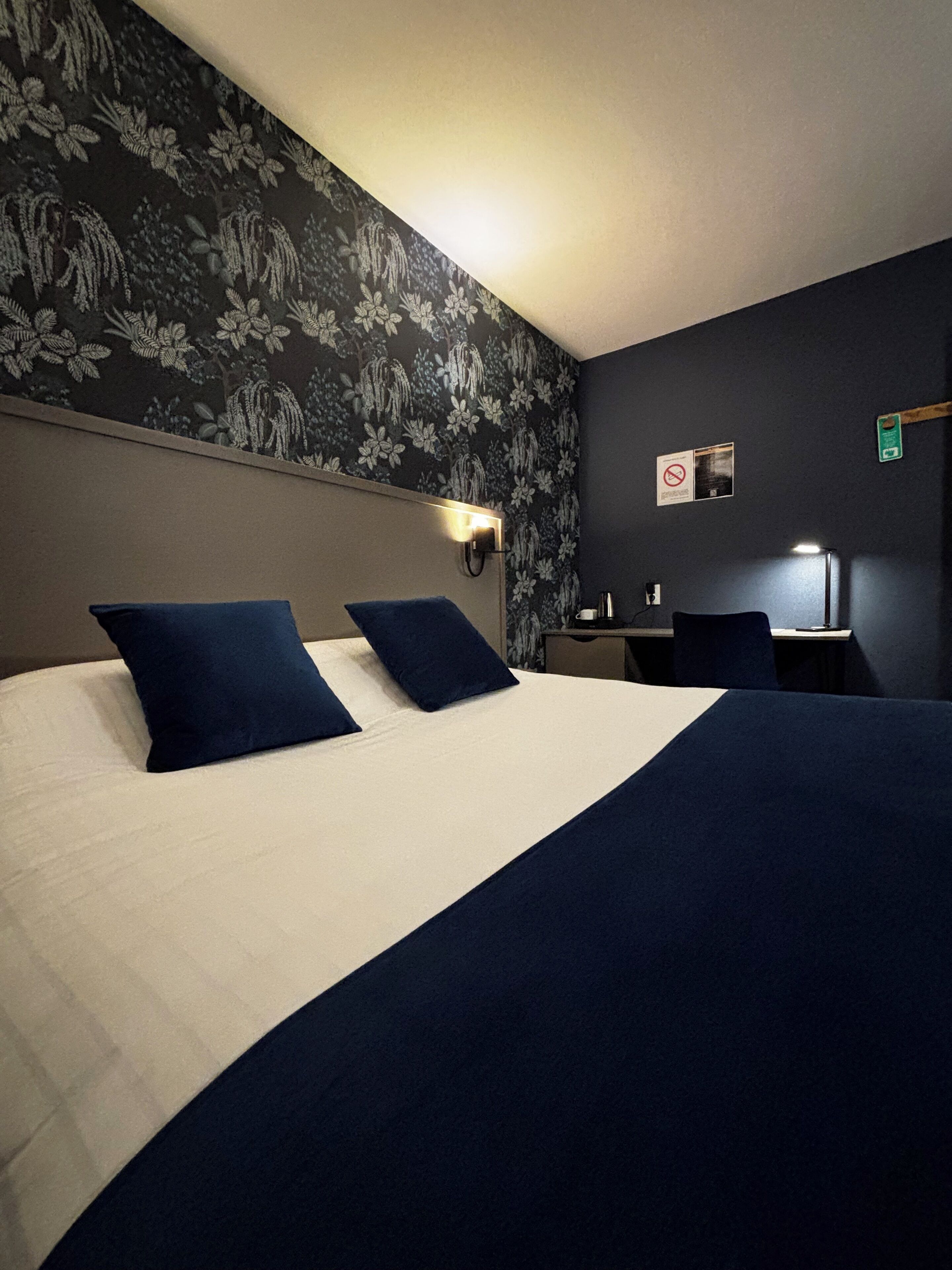 A hotel room with a large bed, two pillows, a desk with a chair, a wall-mounted light, and a floral-patterned wallpaper.